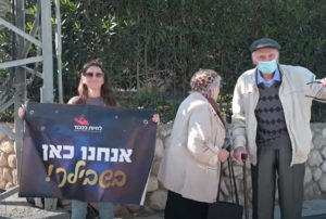 שף של כבוד פרק מסכם - מתנדבת של לחיות בכבוד מקבלת את פני הניצולים עם שלט שכתוב עליו "אנחנו כאן בשבילך!"