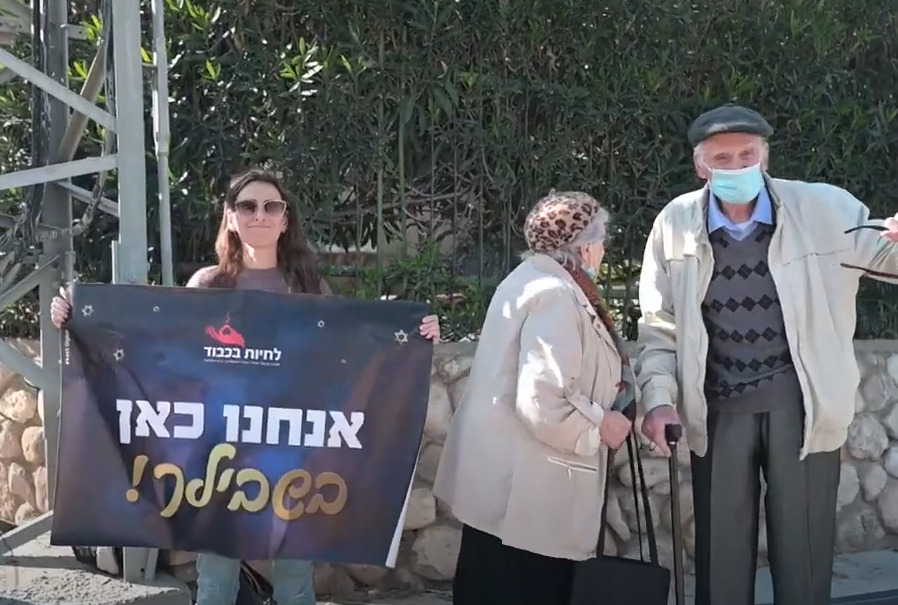 שף של כבוד פרק מסכם - מתנדבת של לחיות בכבוד מקבלת את פני הניצולים עם שלט שכתוב עליו "אנחנו כאן בשבילך!"